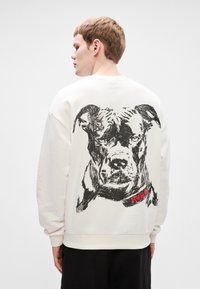 Λευκή μπλούζα φούτερ με ένα μεγάλο μαύρο σκίτσο ενός σκύλου στην πλάτη, με μια κόκκινη ετικέτα "HUGO" γύρω από τον λαιμό του. Μαλακό ύφασμα, χαλαρή εφαρμογή.