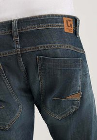 Jeans de mezclilla oscura con un acabado texturizado, que cuentan con múltiples bolsillos y un parche de cuero marrón con el logo en la parte trasera de la cinturilla.