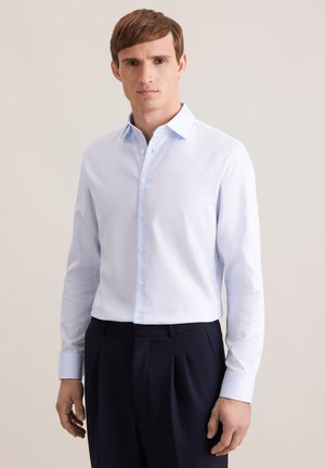 Młody mężczyzna ubrany w jasnoniebieską koszulę zapinaną na guziki i ciemne plisowane spodnie, stojący na tle jednolitego beżowego tła.