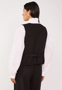 Zwarte wollen vest met een passende uitstraling en een strikdetail op de rug. Draag het over een witte blouse met pofmouwen, die een gestructureerde afwerking laat zien.