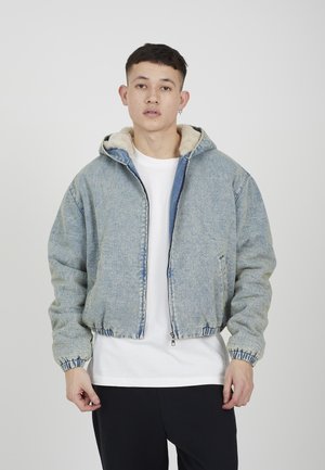 Młody mężczyzna ubrany w jasnoniebieską kurtkę dżinsową z beżowym kapturem wyłożonym polarem, biała koszulka, czarne spodnie, stoi na szarym tle.