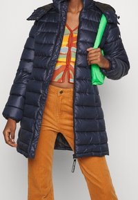 Mörkblå puffjacka med glansig finish, dragkedjeslutning och avtagbar huva. Kombinerad med orange kordbyxor och en färgglad stickad topp.