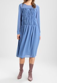 Robe bleue à manches longues avec une texture froncée au niveau du corsage, jupe fluide et longueur mi-mollet. Portée avec des bottes à talons rose clair.