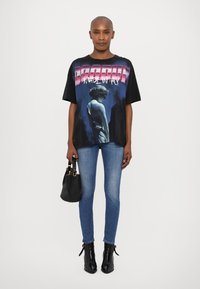Camiseta negra de gran tamaño con estampado gráfico que presenta una estatua clásica, combinada con jeans ajustados azules y botas de tacón negras hasta el tobillo.