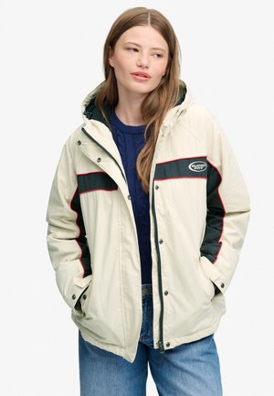 Ung kvinde, der har en creme- og mørkegrøn hættetrøje med rødt kantbånd på, blå sweater og blå jeans på, og kigger til siden med hænderne i lommerne.