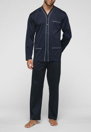 Uomo in piedi a piedi nudi che indossa un completo pigiama blu navy con bordi bianchi, comprendente una parte superiore con bottoni e tasche e pantaloni coordinati.