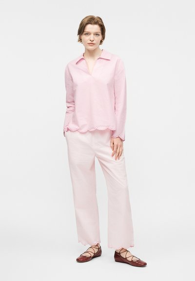 Γυναίκα που φοράει ανοιχτό ροζ μπλούζα και παντελόνια με κυματιστές άκρες, συνδυασμένα με καφέ επίπεδα παπούτσια με κορδόνια, στέκεται μπροστά σε λευκό φόντο.