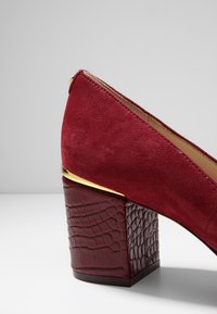Chaussure à talon épais en daim rouge avec talon texturé à motif croco et accent doré sur la partie supérieure du talon, sur un fond gris uni.