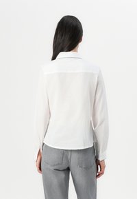 Mujer con cabello largo y negro que lleva una blusa blanca con cuello y jeans grises, de espaldas contra un fondo blanco liso.