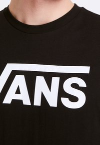 Musta t-paita, jossa on pyöreä pääntie ja rintakehällä rohkea valkoinen "VANS"-logo. Kankaan pinta näyttää sileältä ja kevyesti teksturoidulta.