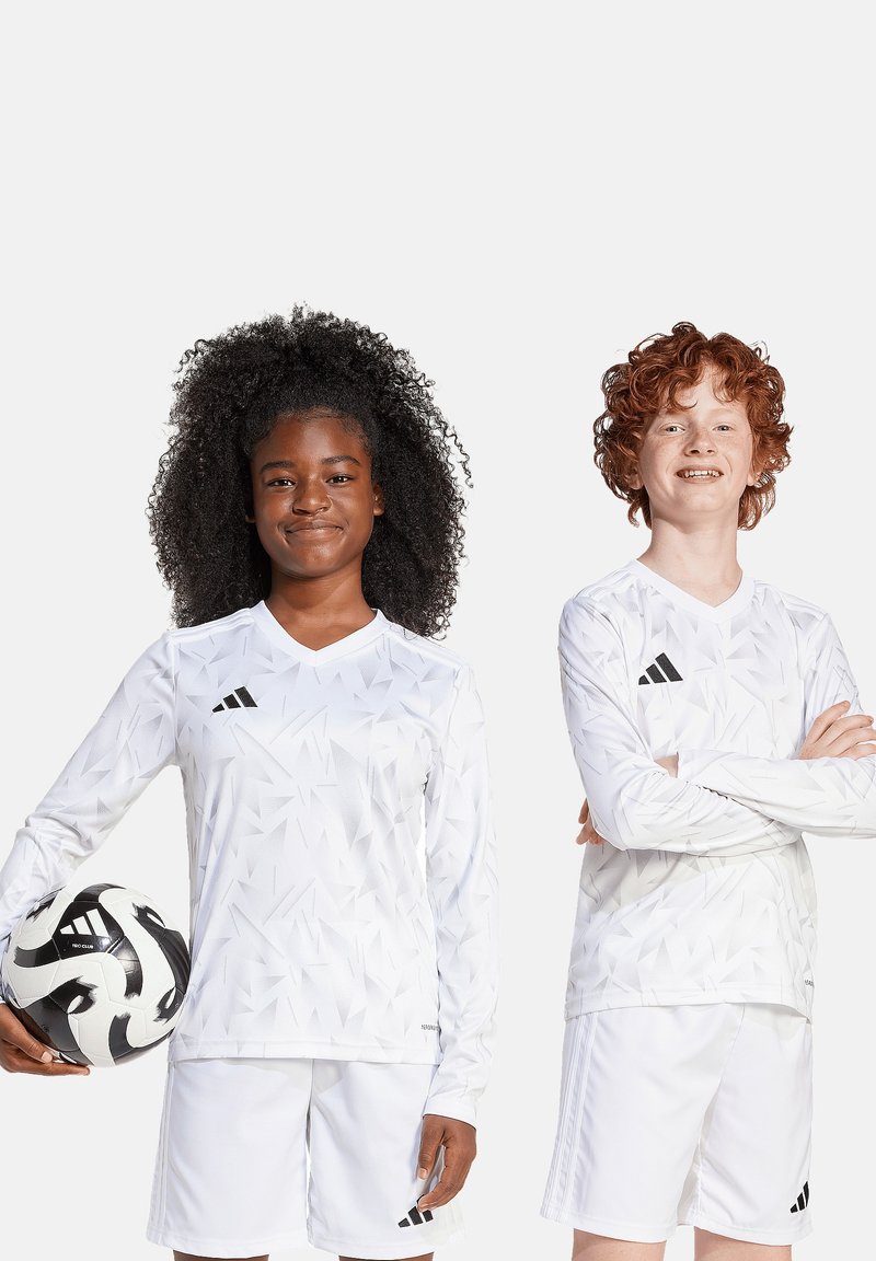Witte voetbalshirts met een geometrisch patroon, zwart Adidas-logo, gecombineerd met witte shorts. Eén speler houdt een zwart-wit voetbal vast.