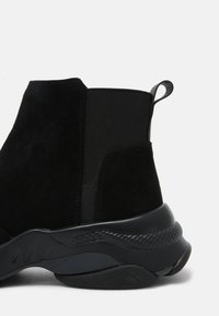 Svart ankelsko med överdel i mocka, elastiska sidopaneler och en texturerad gummisula. Har en dragflik för enkelt påtagande.