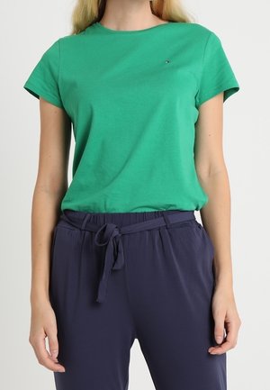 Femme portant un t-shirt vert à manches courtes et un pantalon bleu marine avec une ceinture nouée à la taille, debout devant un fond blanc.