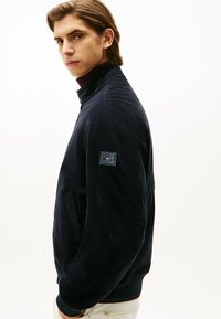 Chaqueta bomber azul marino con una textura suave, cuello alto, bolsillos laterales y un parche de logo cuadrado en la manga izquierda.