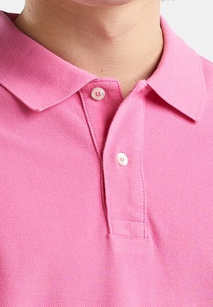 Primer plano de una persona que lleva una camisa tipo polo rosa con cuello abotonada con dos botones blancos abrochados en el escote.