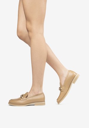 Mocassins en cuir tan avec un petit talon empilé, présentant un accent métallique décoratif et des côtés ouverts pour un design facile à enfiler. Texture lisse.