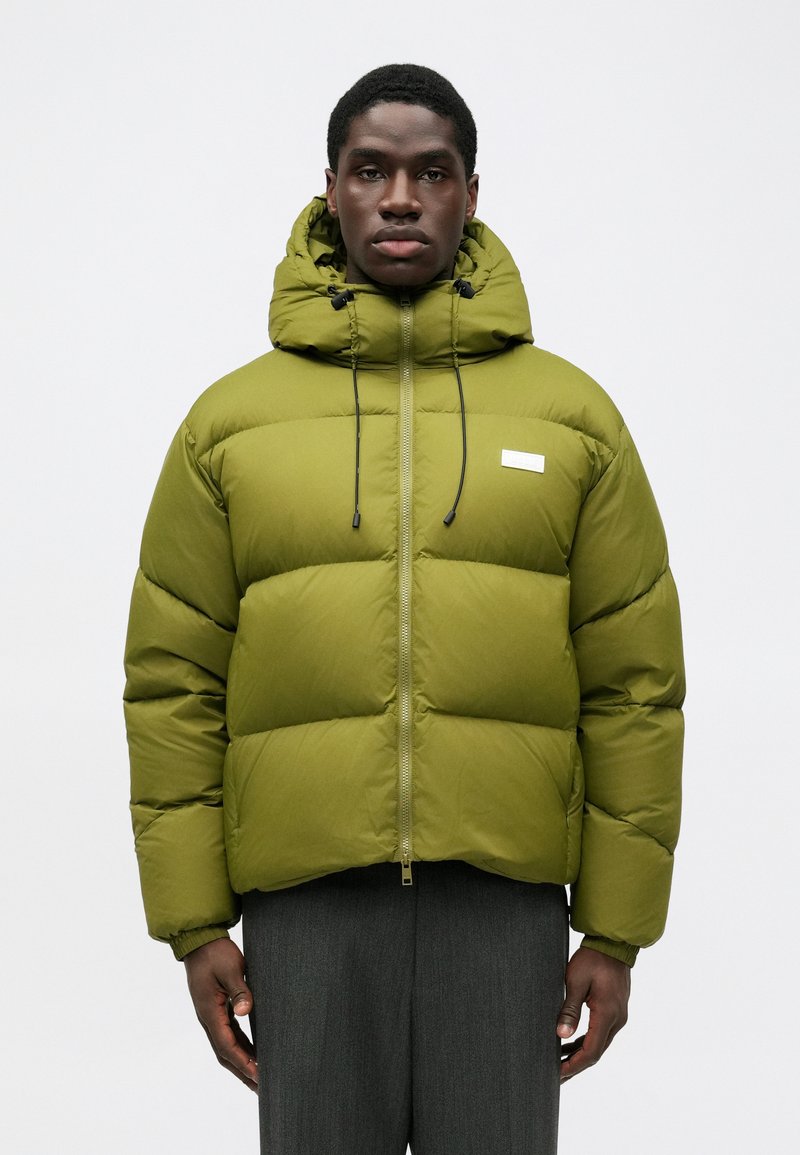 Giacca a vento verde con cappuccio, dotata di chiusura con zip, cordini regolabili e una toppa logo sul davanti. Texture morbida e design trapuntato.