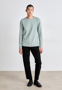 Lichtgroene langslijfshirt met ribbelstructuur, ronde halslijn en geborduurd logo, gecombineerd met zwarte broek en zwarte instap-schoenen.