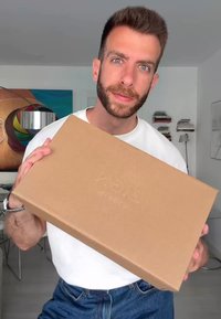 Hnedá kartónová krabica, ktorú drží osoba, s jemne embosovaným logom. Krabica je obdĺžnikového tvaru, s hladkým povrchom a čistými okrajmi.