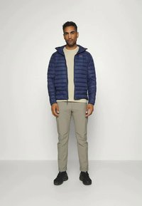 Navy puffer jacket with quilted design, zip closure, and high collar, worn over a light beige top and olive green pants. Black shoes.