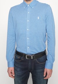 Ljusblå långärmad skjorta med krage, med vita knappar och en broderad logotyp på bröstet, i kombination med mörka jeans och ett svart bälte.