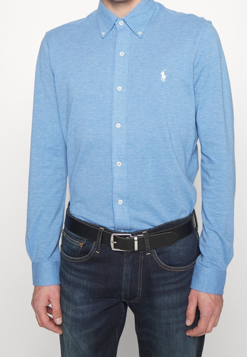 Ljusblå långärmad skjorta med krage, med vita knappar och en broderad logotyp på bröstet, i kombination med mörka jeans och ett svart bälte.