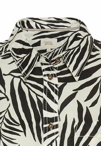 Zwart-witte collshirt met een tropisch bladerenpatroon, knopen en contrasterende stiksels op lichte stof.