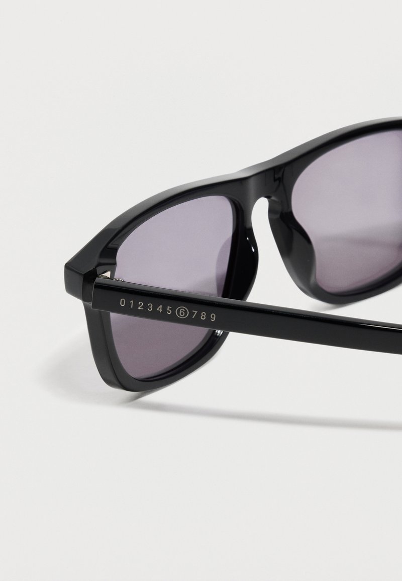 Schwarz gerahmte Sonnenbrille mit dunkel getönten Gläsern, auf einem Bügel ist die Zahlenfolge 012345⑥789 auf weißem Hintergrund aufgedruckt.