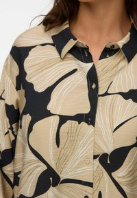 Chemise col berthe présentant une base beige avec des motifs floraux noirs et blancs. Des boutons sur le devant ajoutent du détail au design.
