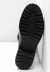 Semelle en caoutchouc noir avec une profonde bande de roulement et un motif en losange, marquée de la taille 37, inclinée sur un fond blanc.