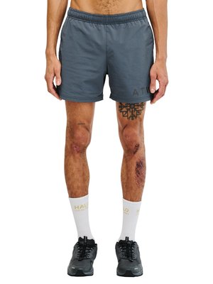 Man draagt grijze korte broek, witte sokken met gele tekst, zwarte sneakers, en heeft tatoeages en blauwe plekken op zijn benen.