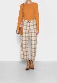 Maglione arancione a maniche lunghe, pantaloni a quadri beige a vita alta e stivaletti marroni alla caviglia. L'outfit include una borsa a tracolla marrone e motivi a quadri.