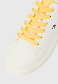 Sneakers bianche con lacci gialli, dettagli cuciti e un piccolo logo "H" rosso, bianco e blu sul lato.