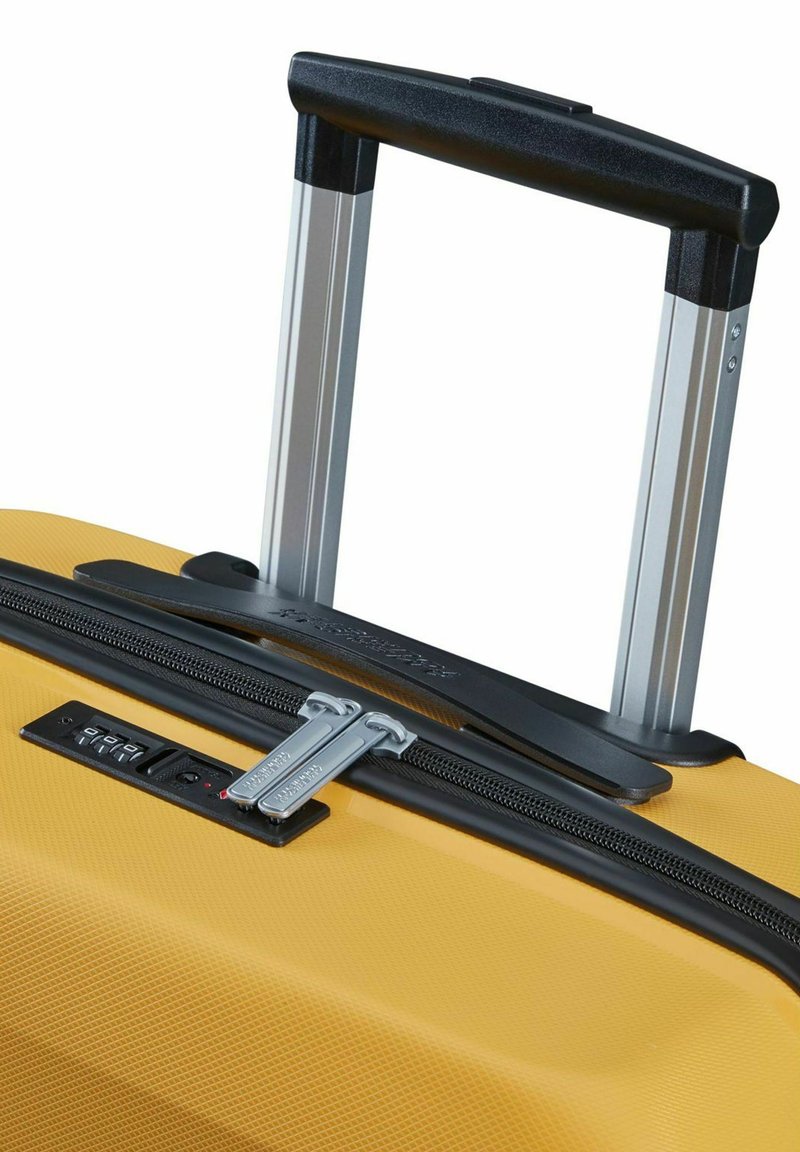 Gele harde koffer met een gestructureerd oppervlak, zilveren telescoophandvat, dubbele ritsen en een combinatieslot aan de voorkant.