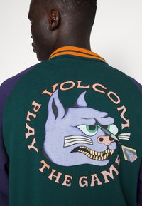 Le sweat-shirt vert et violet présente un grand graphique au dos d'un chat de dessin animé avec des yeux verts et des dents pointues, le texte indique "VOLCOM JOUER AU JEU."