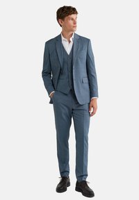 Traje de tres piezas para hombre en tela azul claro con un patrón sutil. Cuenta con un blazer y un chaleco a medida con botones marrones, combinados con zapatos oscuros.