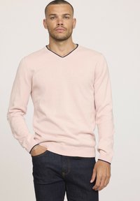 Pull en V rose clair à manches longues, avec un bord bleu marine au niveau du col. Porté sur un jean bleu foncé.