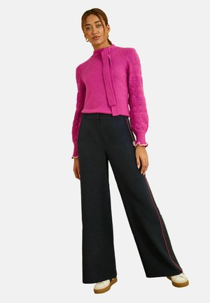 Nő, aki fukszia színű, texturált pulóvert visel puffos ujjakkal, fekete, széles szárú nadrággal, rózsaszín oldalsávval és fehér sportcipővel.