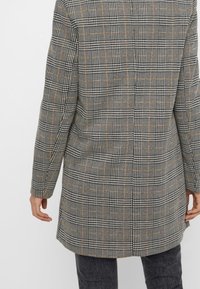 Šedý kostkovaný kabát s strukturovaným designem, s černými a oranžovými akcenty, a hladkým texturovaným materiálem sahajícím do poloviny stehna.