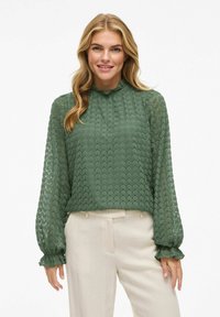 Groene kant blouse met een gestructureerd, ruitvormig patroon, hoge rimpelige kraag, en lange mouwen met rimpelige manchetten, gecombineerd met beige broeken.