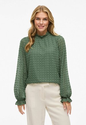 Groene kant blouse met een gestructureerd, ruitvormig patroon, hoge rimpelige kraag, en lange mouwen met rimpelige manchetten, gecombineerd met beige broeken.
