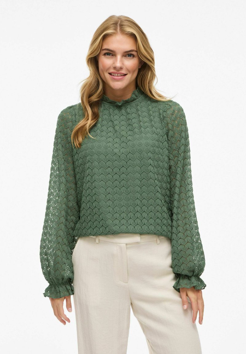 Groene kant blouse met een gestructureerd, ruitvormig patroon, hoge rimpelige kraag, en lange mouwen met rimpelige manchetten, gecombineerd met beige broeken.