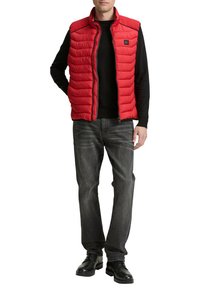 Rote Steppweste mit gesteppter Optik, hohem Kragen und Reißverschluss, getragen über einem schwarzen Langarmshirt, kombiniert mit dunklen Jeans und schwarzen Schuhen.