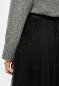 Μαύρη φούστα από δαντέλα με υφή, σε στρώσεις πάνω από διαφανές ύφασμα. Συνδυασμένη με γκρι πλεκτό πουλόβερ που έχει ριμπ μανσέτα.