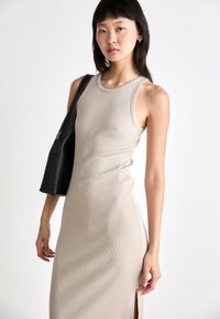 Rochie tricotată ribbed de culoare bej, cu o siluetă ajustată și slit lateral. Asortată cu o geantă de umăr neagră, cu fermoar frontal și accente texturate.