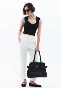 Top corto negro ajustado combinado con pantalones blancos de cintura alta. La modelo lleva un bolso negro estructurado con detalles de cinturón y calza planos negros.