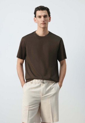 Man staat met zijn handen in de zakken, hij draagt een donkerbruin T-shirt met korte mouwen en licht beige shorts tegen een effen achtergrond.