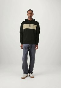 Άνδρας με καπέλο σε μαύρο και μπεζ χρώμα, ξεθωριασμένο γκρι τζιν και λευκά αθλητικά παπούτσια, που στέκεται μπροστά σε απλό φωτεινό φόντο.