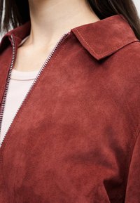 Γυναίκα με σακάκι σουέντ σε σκουρόχρωμη σκιά της σκουριάς, με ασημένιο φερμουάρ, πάνω από ένα ανοιχτό ροζ πουκάμισο, που αφήνει να φαίνεται το γιακά και το πάνω μέρος του στήθους.