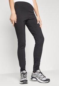 Schwarze Stretch-Leggings mit einer engen Passform, mit seitlichen Nähten und kombiniert mit grau-weißen Wanderschuhen. Weicher Stoff, keine Muster.